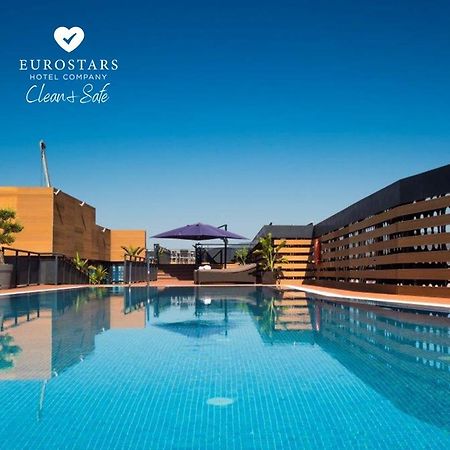 ยูโรสตาร์ส พาเลซ โฮเต็ล Hotel กอร์โดบา ภายนอก รูปภาพ The photo shows an outdoor swimming pool with clear, blue water, reflecting the blue sky above. The pool area is surrounded by modern architectural features, including wooden slats and potted plants. There is also a purple umbrella providing shade. T