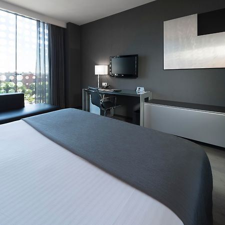ยูโรสตาร์ส พาเลซ โฮเต็ล Hotel กอร์โดบา ภายนอก รูปภาพ The photo shows a modern hotel room featuring a sleek design. In the foreground, there is a bed with a white coverlet and gray accents. To the side, a dark sofa is visible. The room has a large window with sheer curtains, allowing natural light to en