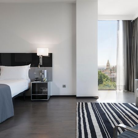 ยูโรสตาร์ส พาเลซ โฮเต็ล Hotel กอร์โดบา ภายนอก รูปภาพ The photo shows a modern hotel room featuring a large bed with white linens and dark cushions, paired with a sleek nightstand. There is a contemporary chair and a small table nearby, along with a striped area rug on the floor. A large window allows n