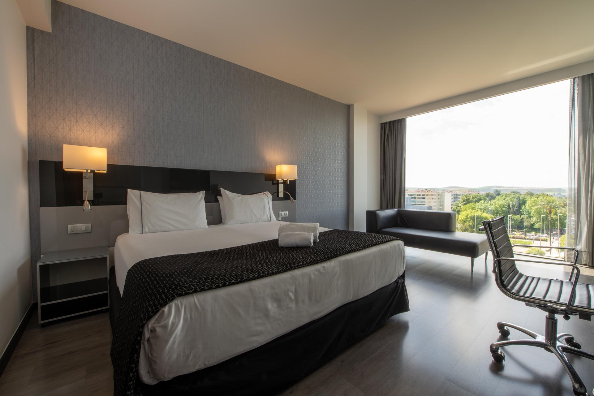 ยูโรสตาร์ส พาเลซ โฮเต็ล Hotel กอร์โดบา ภายนอก รูปภาพ The photo shows a modern hotel room featuring a large bed with white linens and a dark patterned throw. On either side of the bed, there are stylish wall-mounted lamps. A contemporary desk chair is positioned at a small desk, and there’s a sofa near 