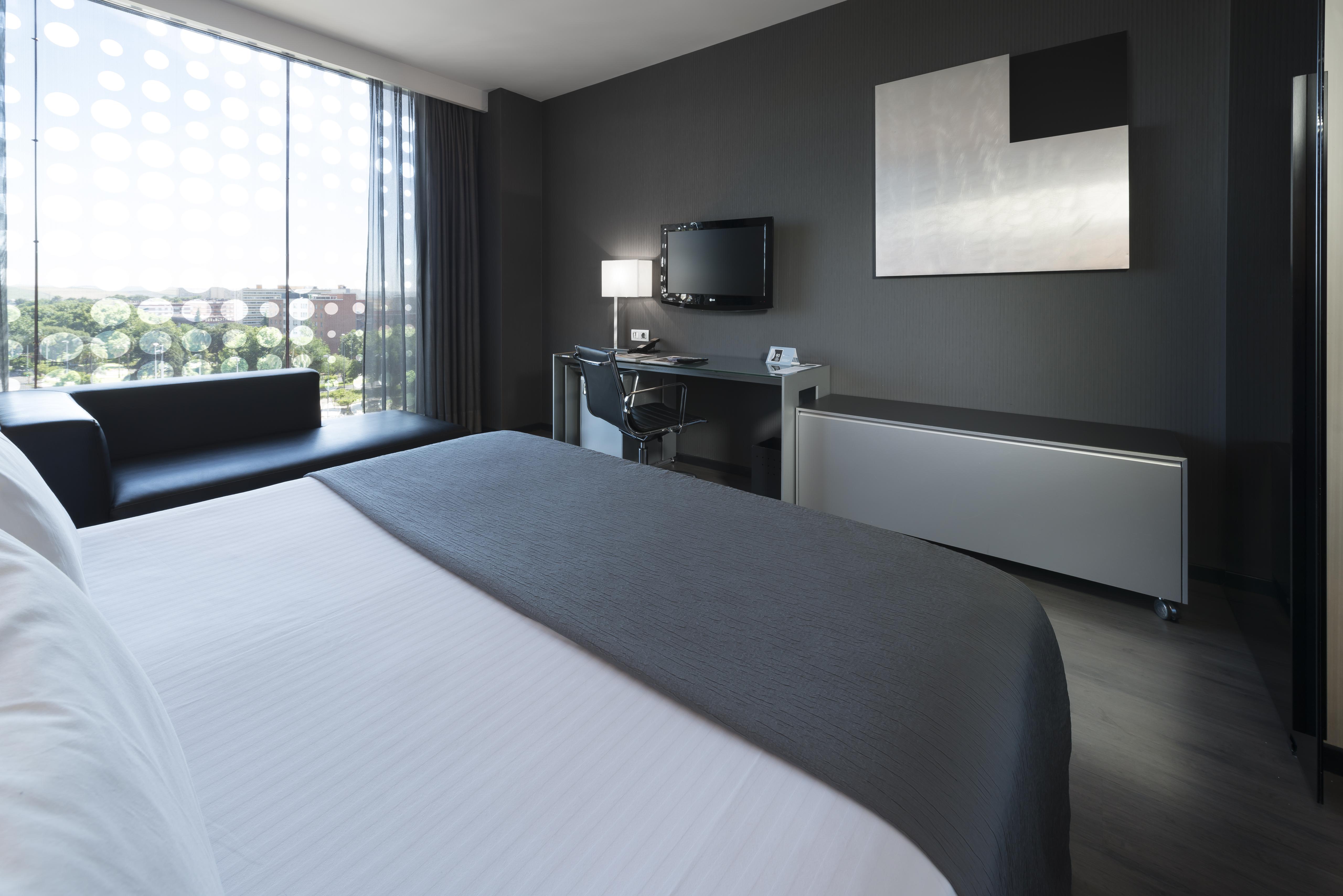 ยูโรสตาร์ส พาเลซ โฮเต็ล Hotel กอร์โดบา ภายนอก รูปภาพ The photo shows a modern hotel room featuring a sleek design. In the foreground, there is a bed with a white coverlet and gray accents. To the side, a dark sofa is visible. The room has a large window with sheer curtains, allowing natural light to en