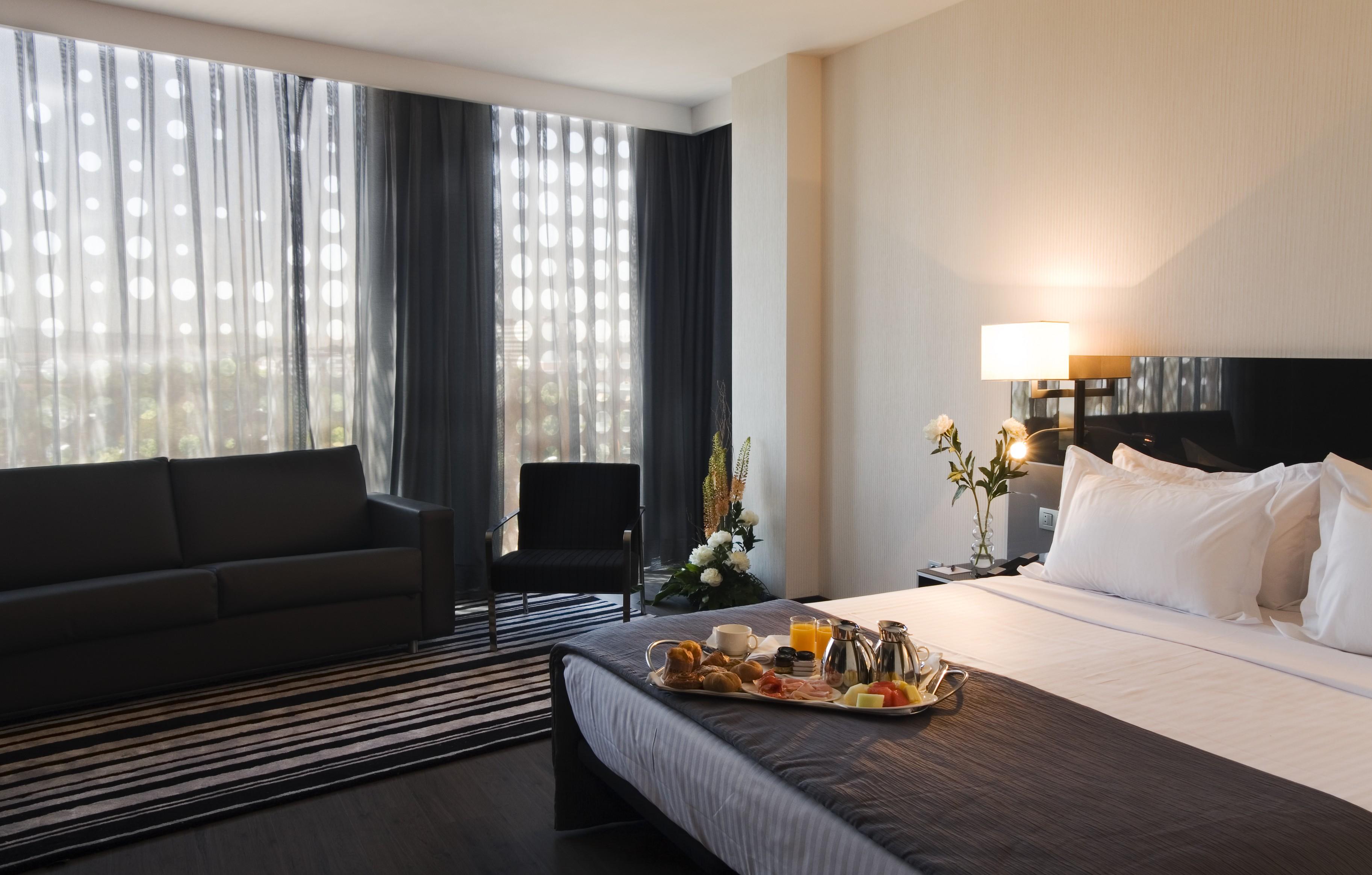 ยูโรสตาร์ส พาเลซ โฮเต็ล Hotel กอร์โดบา ภายนอก รูปภาพ The photo shows a modern hotel room featuring a neatly made bed with white linens and a dark grey blanket. There is a tray on the bed with a breakfast spread, including pastries, fruit, and beverages. To the left, there is a comfortable-looking couch