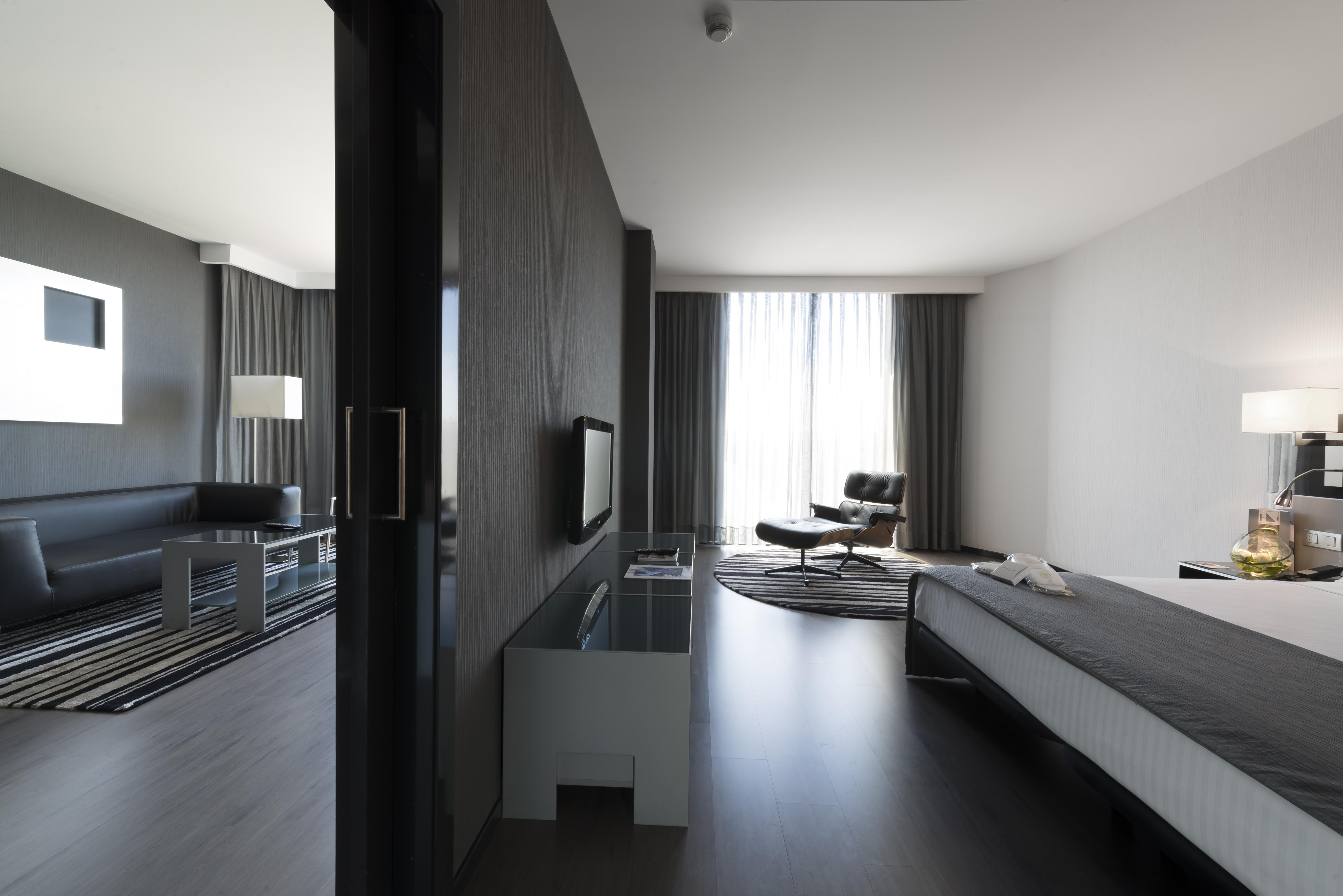 ยูโรสตาร์ส พาเลซ โฮเต็ล Hotel กอร์โดบา ภายนอก รูปภาพ The photo shows a modern hotel room or suite featuring a contemporary design. On the left side, there is a living area with a black sofa, a coffee table, and light fixtures. The large windows, covered with sheer curtains, let in natural light, enhanc