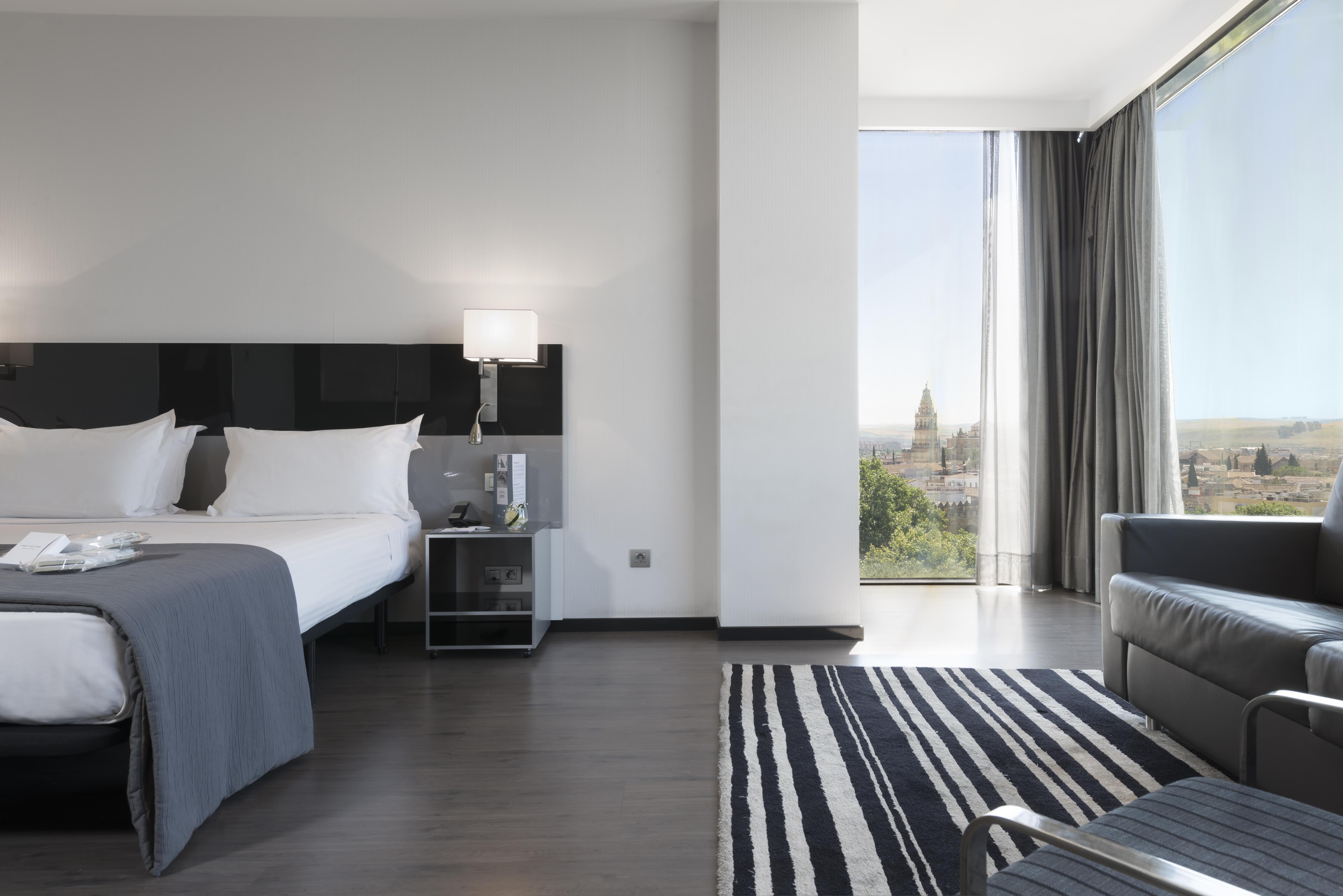 ยูโรสตาร์ส พาเลซ โฮเต็ล Hotel กอร์โดบา ภายนอก รูปภาพ The photo shows a modern hotel room featuring a large bed with white linens and dark cushions, paired with a sleek nightstand. There is a contemporary chair and a small table nearby, along with a striped area rug on the floor. A large window allows n
