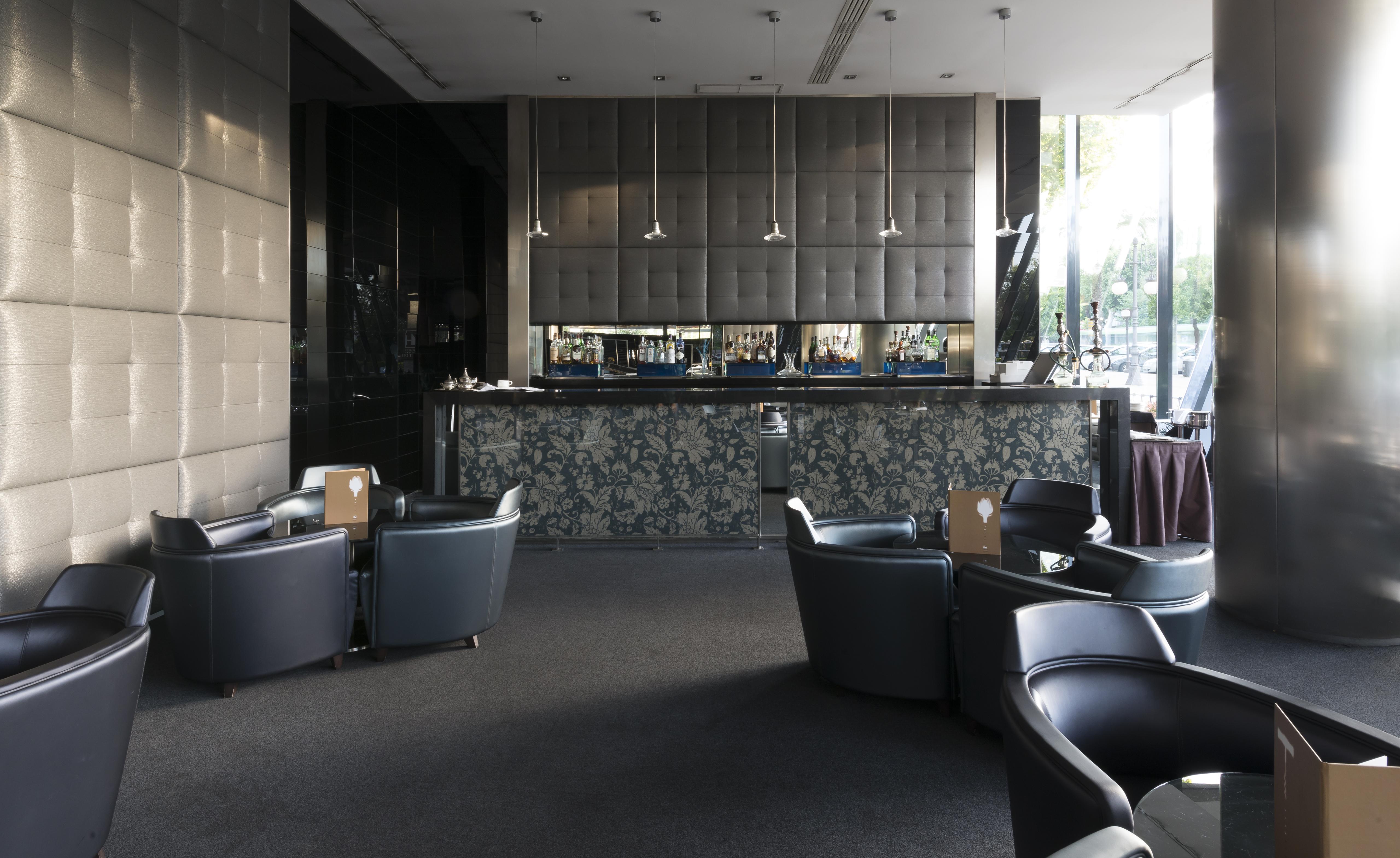 ยูโรสตาร์ส พาเลซ โฮเต็ล Hotel กอร์โดบา ภายนอก รูปภาพ The photo shows a modern bar or lounge area. In the foreground, there are several comfortable black armchairs arranged around small tables. The background features a sleek bar counter with a dark, textured wall behind it. The bar is illuminated with 