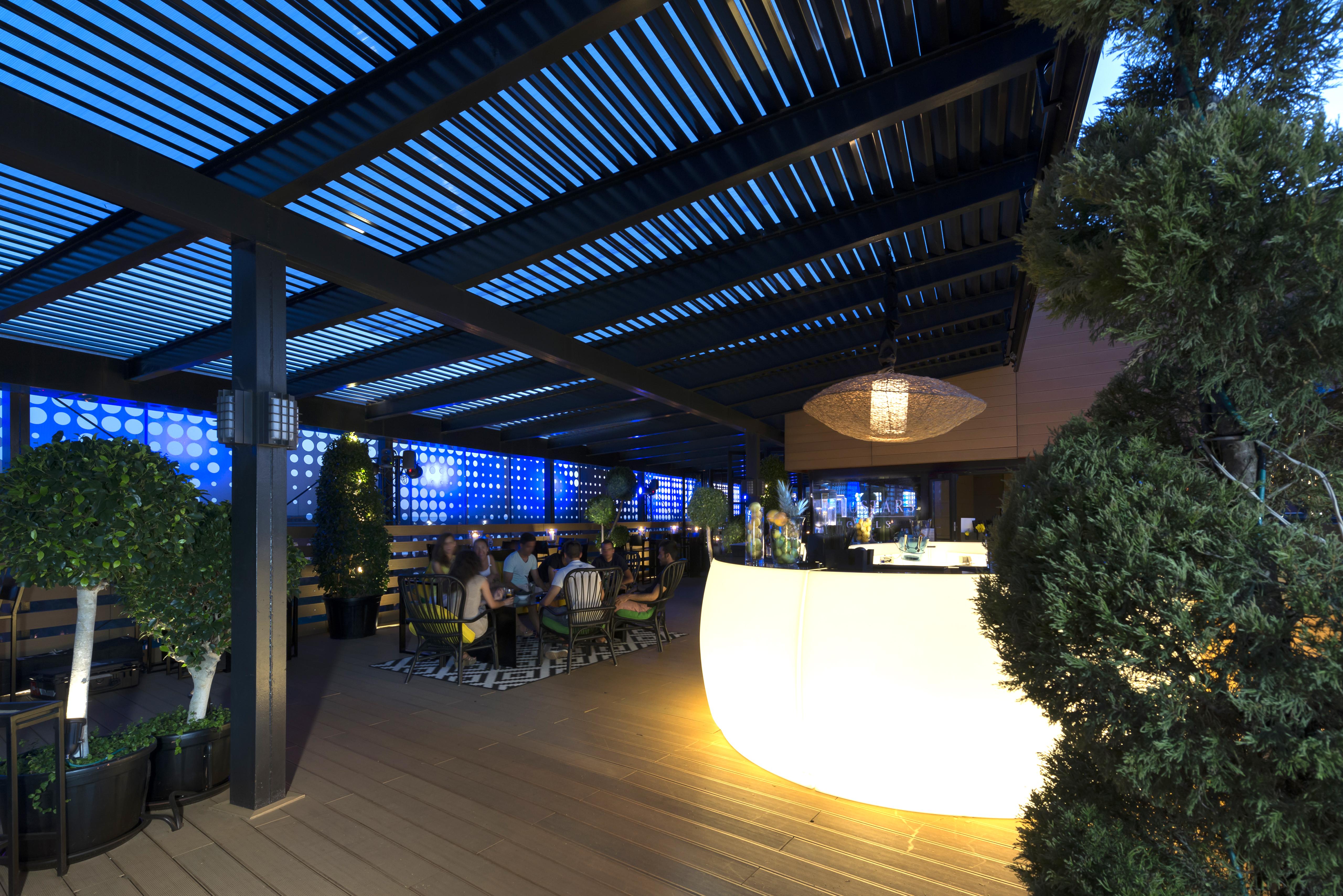 ยูโรสตาร์ส พาเลซ โฮเต็ล Hotel กอร์โดบา ภายนอก รูปภาพ The image shows a modern outdoor bar or restaurant area. The space features a sleek, illuminated bar that stands out with its curved shape. Patrons can be seen seated around tables, enjoying the ambiance. The overhead structure consists of slatted pa