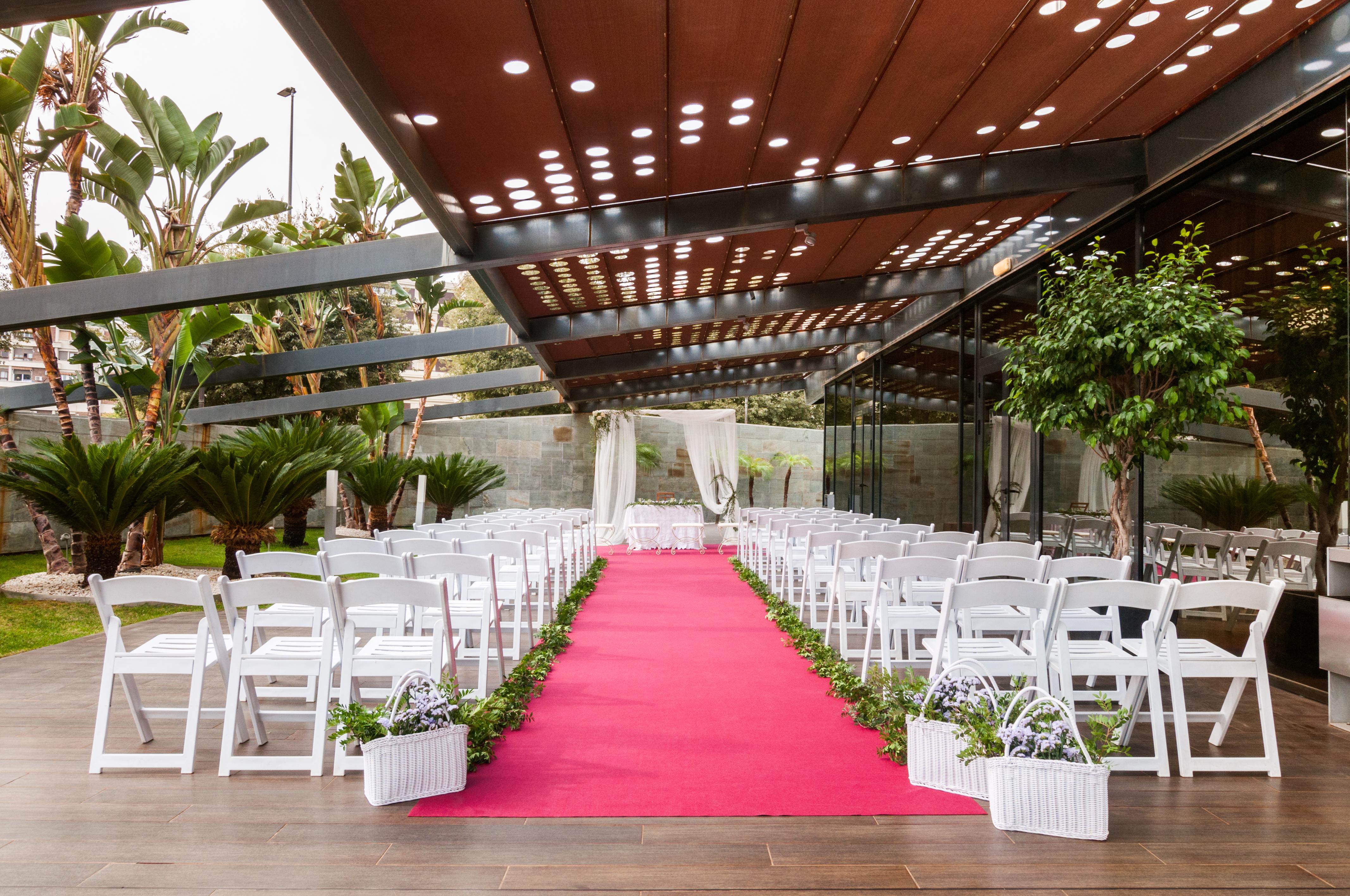 ยูโรสตาร์ส พาเลซ โฮเต็ล Hotel กอร์โดบา ภายนอก รูปภาพ The photo shows a wedding setup in an outdoor venue. There are white chairs arranged in rows on either side of a vibrant pink carpet leading to a floral arch or backdrop at the front. The area is partially shaded by a modern structure with a metal fr