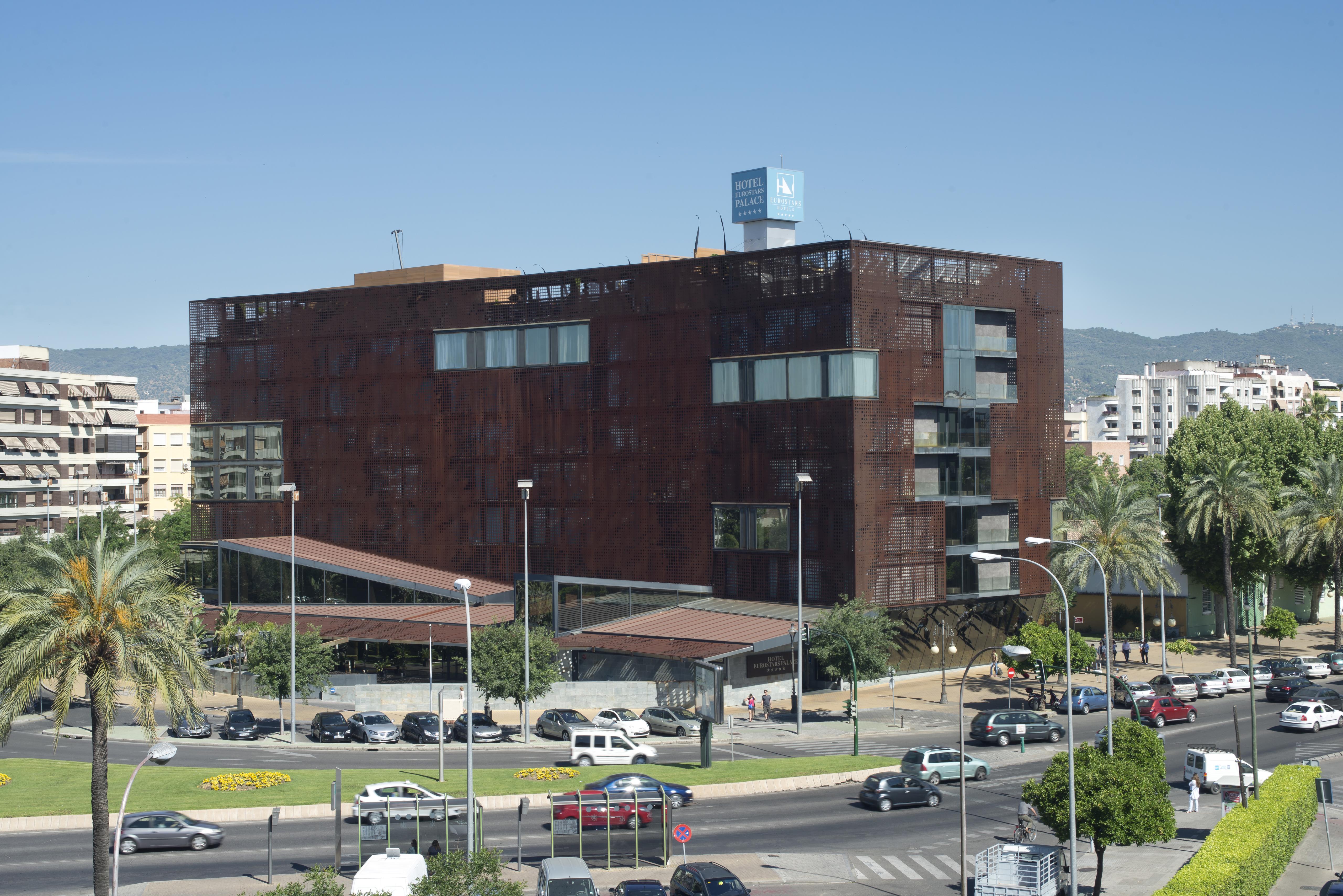 ยูโรสตาร์ส พาเลซ โฮเต็ล Hotel กอร์โดบา ภายนอก รูปภาพ The photo shows a modern building characterized by a distinctive brown, rust-like exterior, which gives it an industrial appearance. The structure has multiple large windows, allowing for natural light to enter. There are several palm trees surroundi
