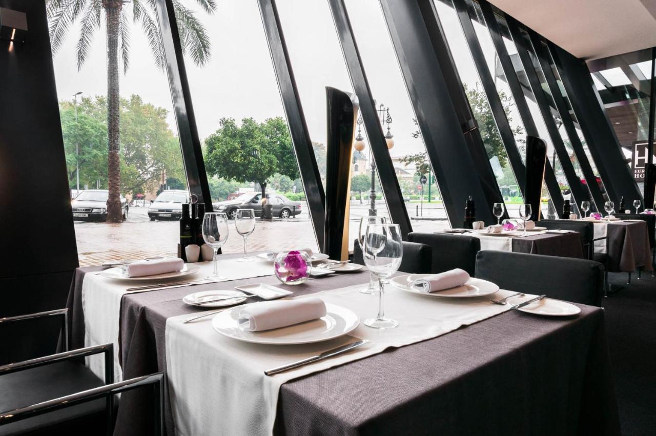 ยูโรสตาร์ส พาเลซ โฮเต็ล Hotel กอร์โดบา ภายนอก รูปภาพ The photo shows an elegant restaurant interior with large windows featuring modern angular designs. There are two dining tables set up, each covered with white and dark tablecloths. The tables are elegantly arranged with plates, cutlery, and wine gla